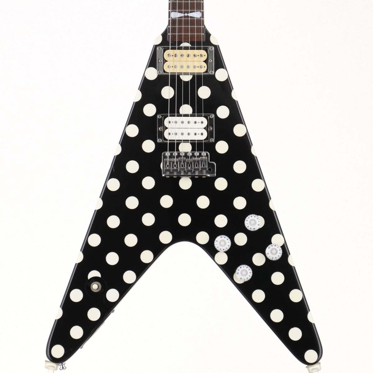 The body of a black Flying V electric guitar covered in large cream polka dots, shot straight on against a white background, with two humbucking pickups, a tremolo bridge and three control knobs