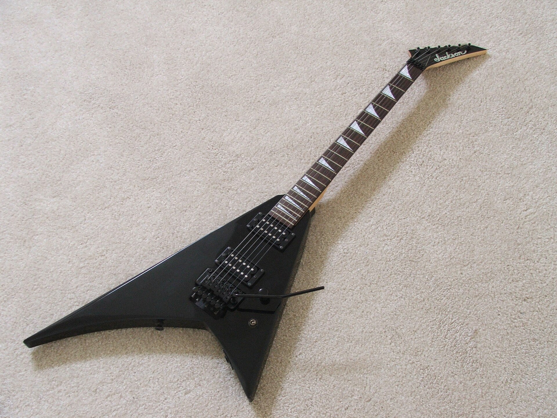 A black Jackson Randy Rhoads guitar lying on pale carpet, showing the asymmetrical V body with one wing shorter than the other, shark fin inlays and a Floyd Rose tremolo