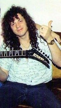 Zeke Clark as a young man with long curly hair, seated on a couch holding a Kramer guitar and giving a thumbs up