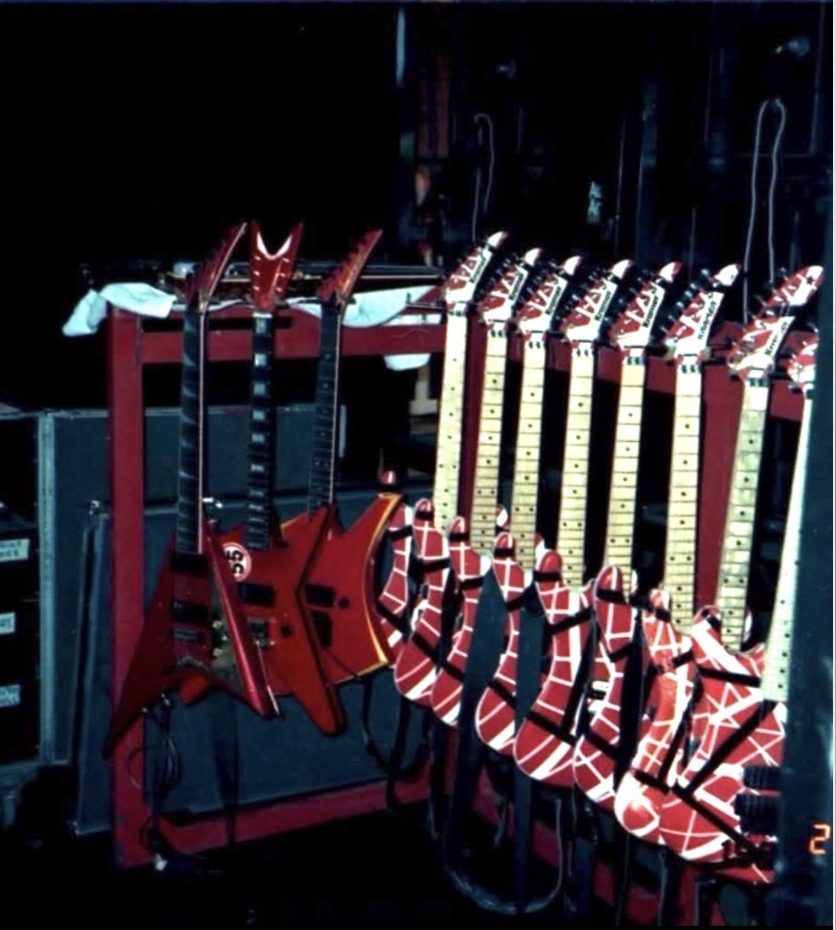 A period colour photograph of Eddie Van Halen's backstage guitar rack: a row of red, white and black striped Kramers hanging by their necks, with red pointed-body guitars on a stand alongside and flight cases behind