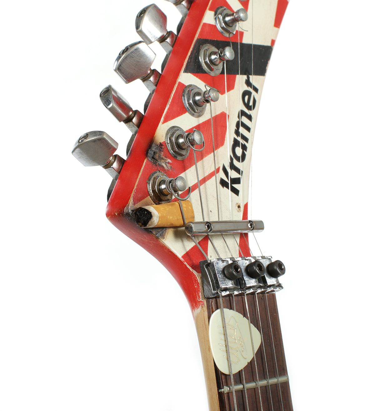 Close detail of the Kramer headstock: a cigarette butt wedged between the strings and the nut, a burn mark in the finish beside it, and a signed pick tucked under the strings on the fingerboard