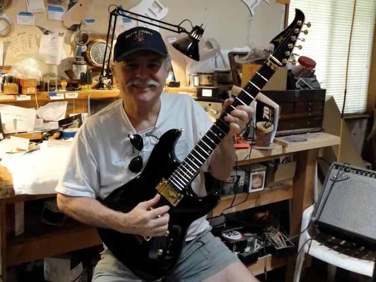 Wayne Charvel sitting in his workshop holding a black electric guitar with gold hardware, surrounded by tools and parts bins